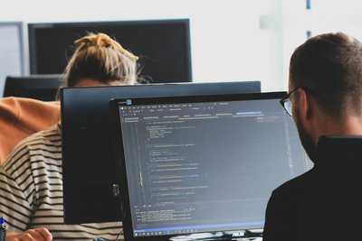 Two professionals discussing code on multiple computer screens in an office.